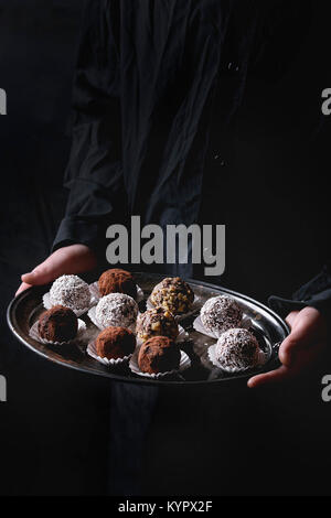 Variety of homemade dark chocolate truffles with cocoa powder, coconut, walnuts on vintage tray in kid's hands in black shirt. Dark background. Stock Photo