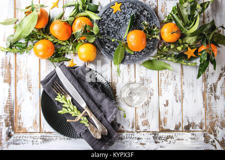 Christmas table decorations with clementines or tangerines with leaves and green branches on black ornate board. Empty plate with cutlery and textile Stock Photo