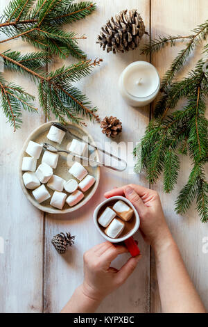 Hands holding a cup of hot chocolate,white candle,spruce cones and spruce tree branches on the table Stock Photo