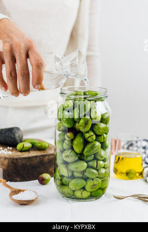 Woman pouring water on green olives in a glass jar. A cup of olive oil and a wooden mallet accompany. Stock Photo