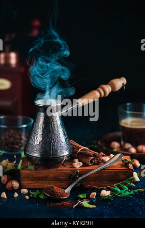 Steaming cezve with hot coffee on dark background with beans, cinnamon, nuts, leaves and coffee grinder. Brewing coffee concept. Stock Photo