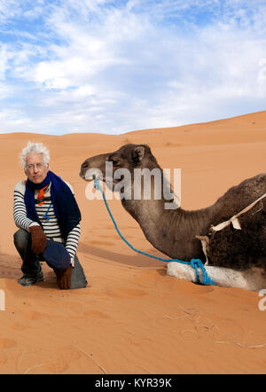 Senior man on an adventure travel trip to Morocco. Shown on the sand with a camel in the Sahara Desert. Theme is active retirement. Stock Photo