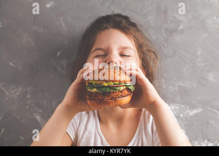 The little girl is eating a healthy baked sweet potato burger with a whole grains bun, guacamole, vegan mayonnaise and vegetables. Child vegan concept Stock Photo