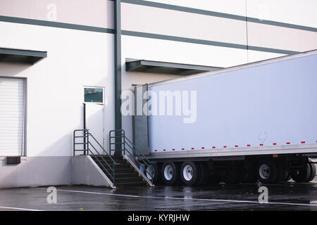 Long dry van semi trailer with dual axles loading and unloading commercial cargo in concrete walls warehouse dock with gates and steps Stock Photo