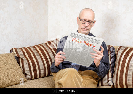 A man wearing glasses is sitting on a couch at home, reading a newspaper reporting fake news Stock Photo