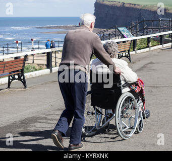 Elderly man pushing edlerly woman in wheelchair on seafront. UK Stock Photo