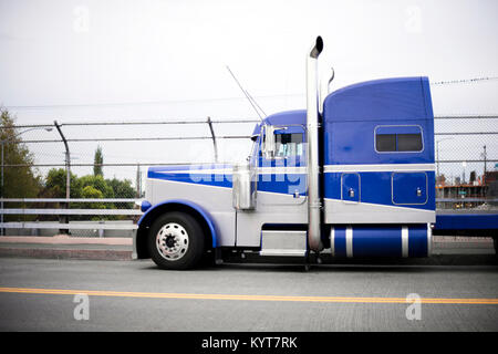 Profile of the blue American classic popular powerful big rig semi truck tractor with chrome accessories and tall exhaust pipes going on the road Stock Photo