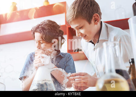 Experimenting in the laboratory. Smart schoolboys both wearing shirts looking at a fuming flask while performing an experiment with different liquids  Stock Photo