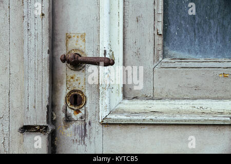 Old vintage and rusty door handle installed on an old grey weathered and cracked grunge wooden door with glass in an abandoned house. Stock Photo