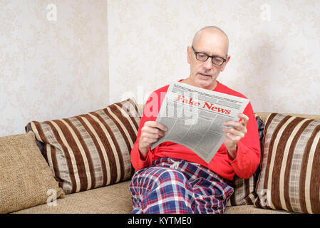 A man wearing glasses and pajamas is sitting on a couch at home, reading a newspaper reporting fake news Stock Photo