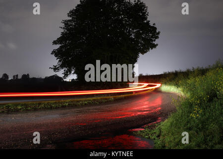 Car light trail around a tree - Long exposure of a car's rear lights bending around a tree in the English countryside. Wet road reflecting a red glow Stock Photo