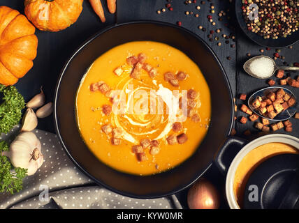 Pumpkin creme soup in a dark crockery served with croutons, crushed nuts and cream, top view. Around are ingredients: pumpkins, carrots, onions, garli Stock Photo