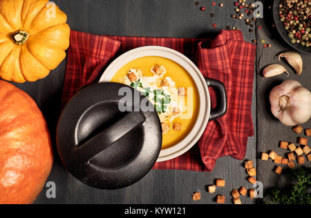 Pumpkin creme soup in a dark ceramic pan served with croutons, crushed nuts, parsley and cream, top view. Around are ingredients for the soup: pumpkin Stock Photo