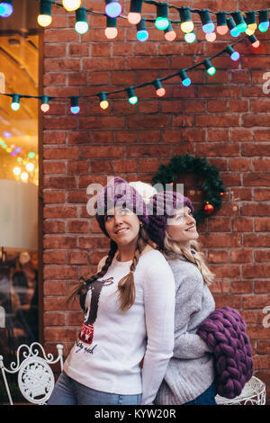 Two women in purple knitted hats, purple infinity scarf and blue jeans make posing with brick wall and Christmas garland on the background Stock Photo