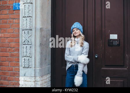 Young blonde woman in blue knitted hat, white fluffy mittens, grey sweater and blue jeans make posing with wooden door on the background Stock Photo