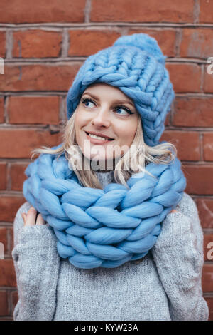 Young blonde woman in blue knitted hat, blue khitted infinity scarf and grey sweater make posing with brick wall on the background Stock Photo