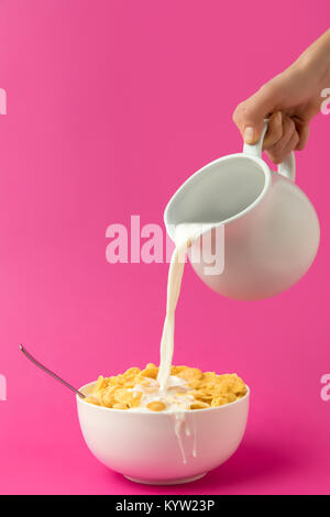 cropped shot of hand holding jug and pouring milk into bowl with corn flakes isolated on pink Stock Photo