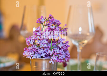 Lilac flowers as spring bouquet in a metal cup on a table setting with wine glasses in the background. Warm shades in a cozy interior.