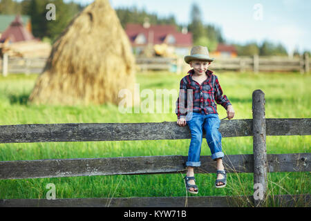 Little boy in hat sitting on fence against of haystack. Child on a hedge against backdrop of rural landscape. Young cowboy smiling shyly and looking a Stock Photo