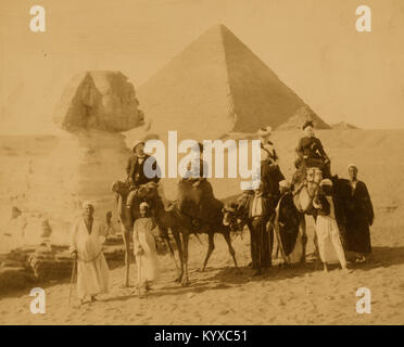 Three women and a man in western attire, seated on camels, several local men stand before the camels holding the reins, a pyramid and the sphinx in background Stock Photo