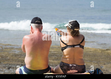 Brirs abroad: woman applying spray suncream to sunburnt back of man on beach in Spain. Stock Photo