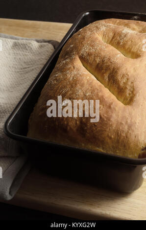 Freshly baked, home made loaf of organic bread in tin on wooden chopping board with folded oven mitt to the side. Stock Photo