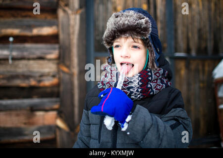 Happy child sticking out a tongue tasting an icicle with wooden cabin in the background. Boy enjoying snow. Stock Photo