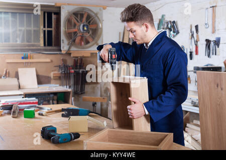 Young positive guy using drilling machine to make drawer at workshop Stock Photo