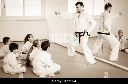 Cheerful young male coach explaining new maneuvers to children in karate class. Focus on the boy Stock Photo