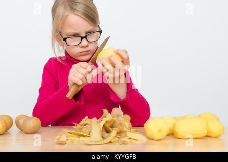 A girl is wearing a red turtleneck and glasses. It peels a potato with a sharp kitchen knife. Before him lies a heap of potato peel Stock Photo