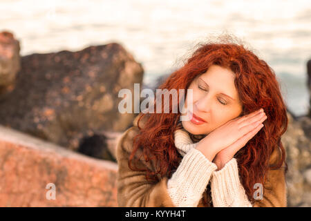 Classy mature woman taking a nap in a winter dress with rocks and sea as background Stock Photo