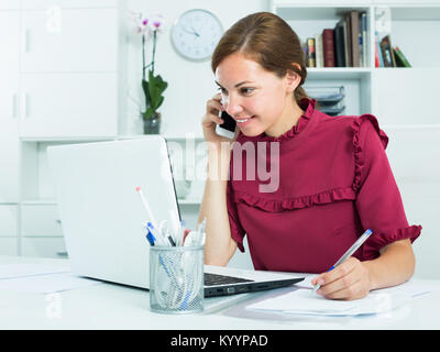 young concentrated female employee answering on phone and taking notes at office Stock Photo