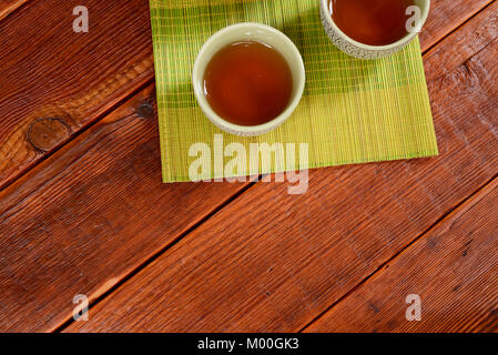 Still life of traditional Chinese crackle glaze tea cups filled with oolong tea on a green bamboo mat on top of dark brown, rough wooden table Stock Photo