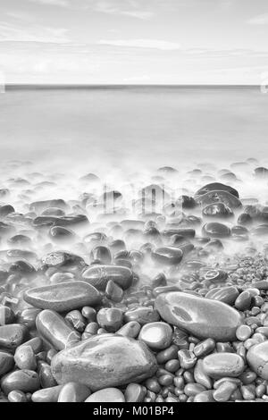 Black and white image of pebbles in blurry water at the shoreline. Stock Photo