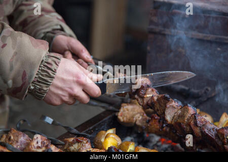 The cook prepares meat on the skewer and tests the readiness of the meat with a large knife. Stock Photo