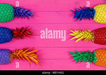 Six brightly colored pineapple tops with leaves in the colors of the spectrum as side borders over a pink wood background with center copy space Stock Photo