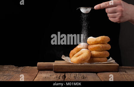 Baker sprinkles sweet donuts with powder sugar on black background. Delicious, but unhealthy food on the old wooden table with copy space. Stock Photo