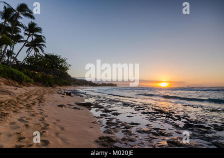 Sunset over Sunset Beach on North Shore of Oahu, Hawaii with waves rolling in over rocks and palm trees in foreground Stock Photo