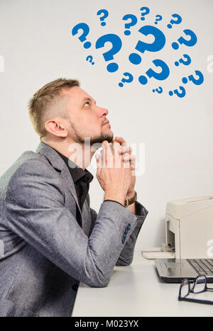 man with chin on hands thoughtfully looking upwards on question marks sitting at table with laptop in office Stock Photo