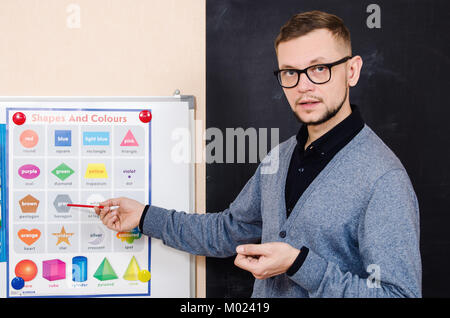 bearded man a teacher at the blackboard explains the shapes and colors on a special poster. grey hexagon. Stock Photo
