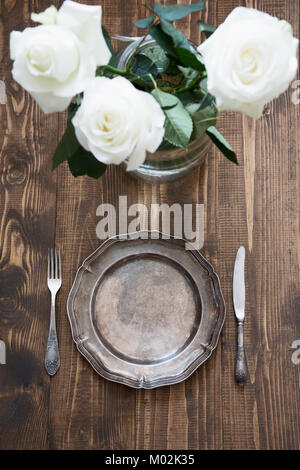 Romantic table setting with white rose, vintage dishware, silverware and decorations on wooden board. Selective focus. Stock Photo