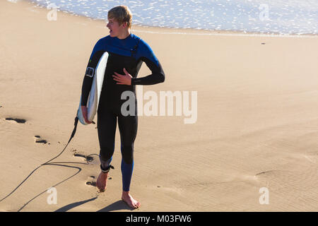 Surfer holding surf board walking along seashore at Bournemouth beach, Bournemouth, Dorset UK in January Stock Photo