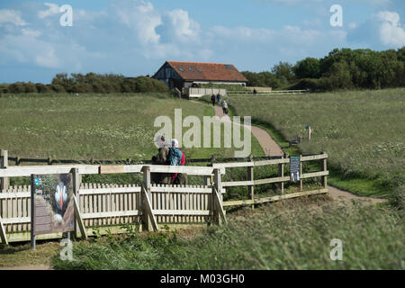 2 visitors to RSPB nature reserve stand at viewing point while other people walk near visitor centre beyond - Bempton Cliffs, Yorkshire, England, UK. Stock Photo