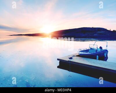 Motorboat for sport fishing tied to a wooden floating dock. Fishing boat with fishing rods in hangers  in bay port, sunset calm water. Stock Photo