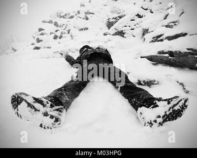 Funny boy laying in snow and makes an angel with spread arms. Kid play game in fresh snow on stony hill. Stock Photo