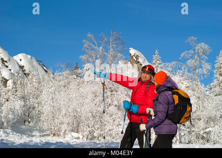 two hikers in winter on a clear frosty day against the backdrop of snowy forest and rocks; man looking at the woman showing a hand in the distance Stock Photo