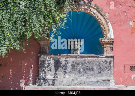 Old stone water basin, with an arch, a blue interior and other decorative elements, a pink stucco wall and green shady tree, in San Miguel de Allende Stock Photo