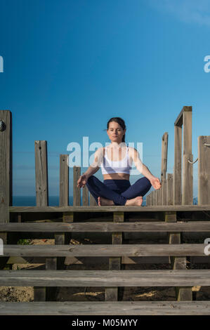 woman meditating in the lotus position sitting on wooden steps by the sea. Stock Photo