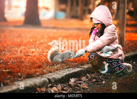 Little girl in winter coat feeding nuts to squirrel in a park in Autumn Stock Photo