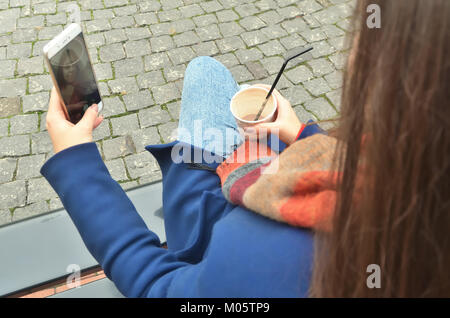 A young attractive woman sitting on the outside and makes selfie on phone. Sit back to the photographer. Holds a cup of coffee in her hand. Stock Photo